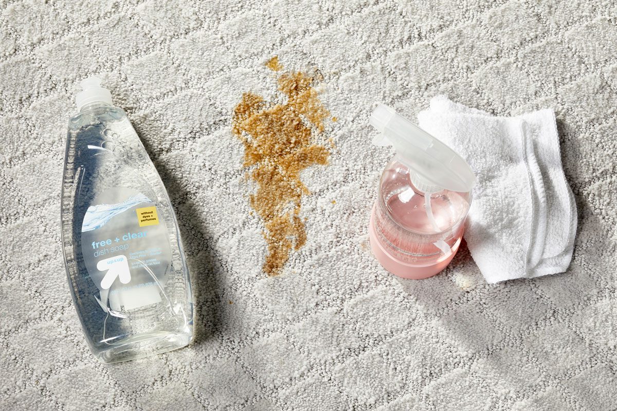 bottle of clear dish soap, spray bottle, and cleaning cloths next to a stain on a light gray carpet for Homemade Dish Soap Carpet Cleaner