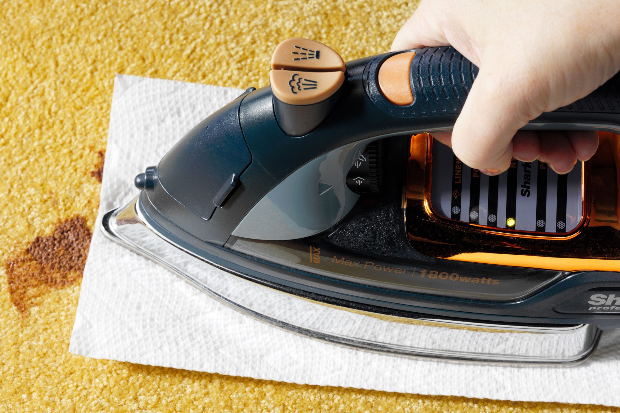 hand using an iron over a paper towel on the chocolate stain to remove it from the yellow carpet
