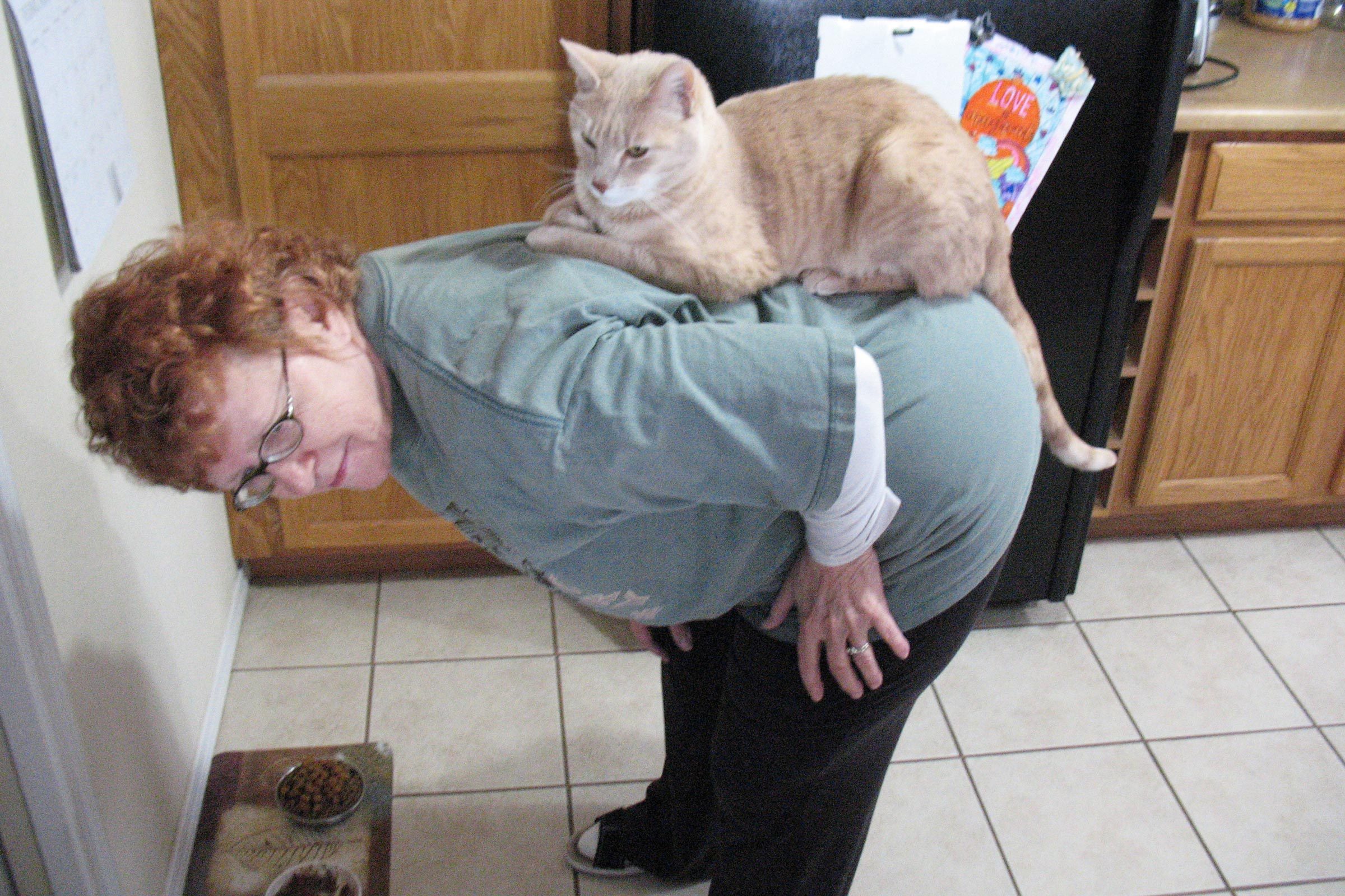 a woman bent over in the kitchen with a cat seated on her back