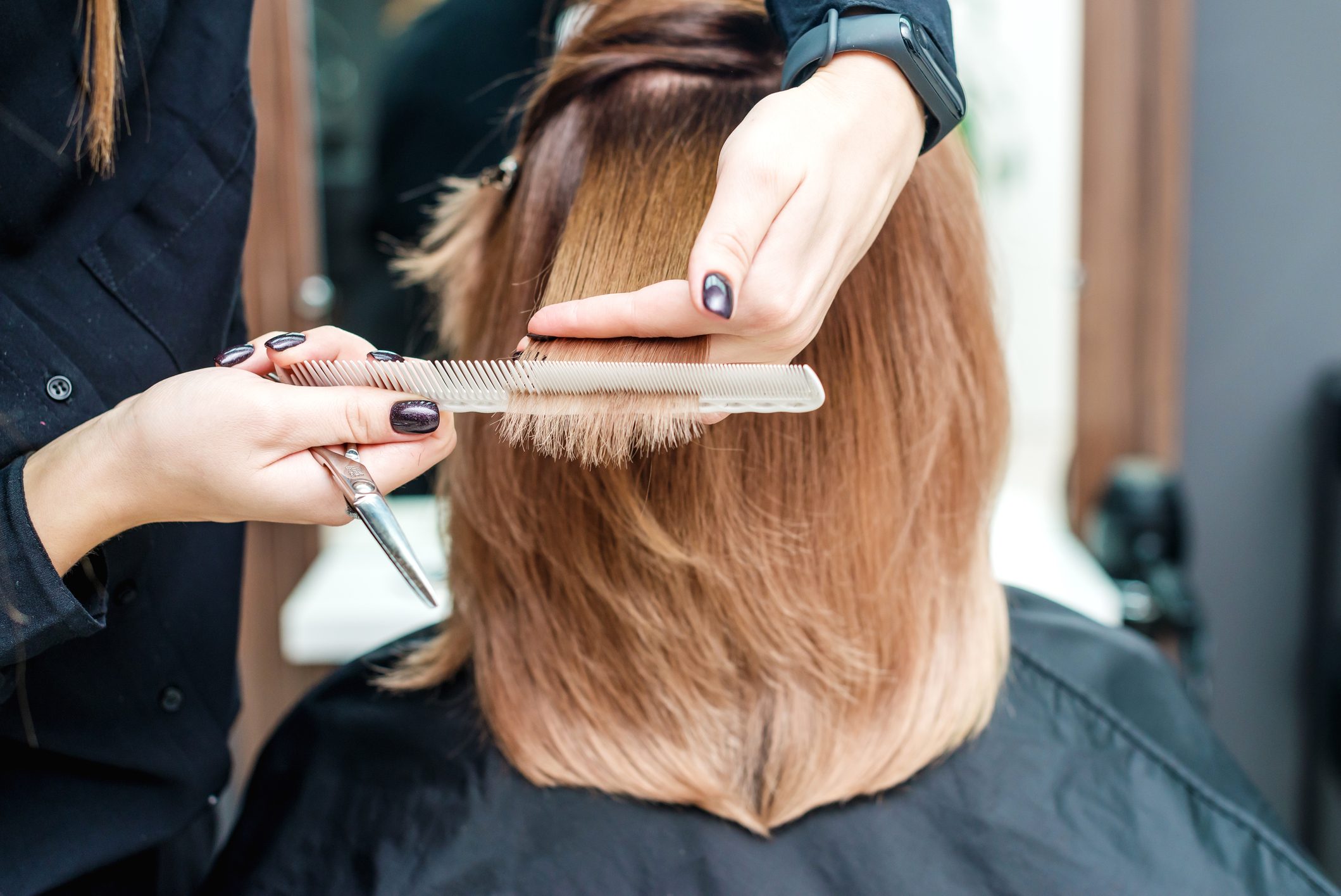 Hands are trimming brown hair with scissors and a comb in a salon setting.