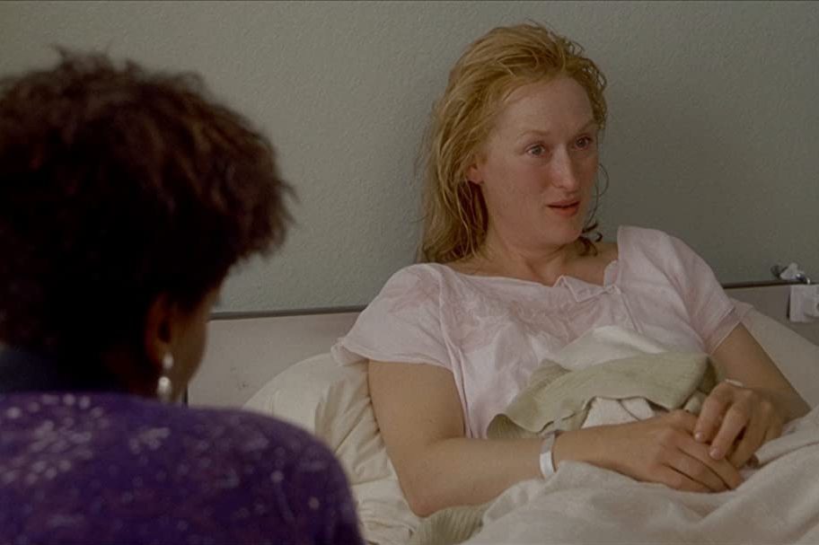 A person reclines in a hospital bed, conversing with someone nearby, in a room with light-colored walls.
