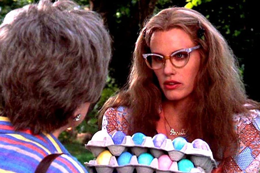 Woman holds carton of colorful eggs, talking to another person, outdoors with trees and greenery in the background. Name necklace reads "Harold."