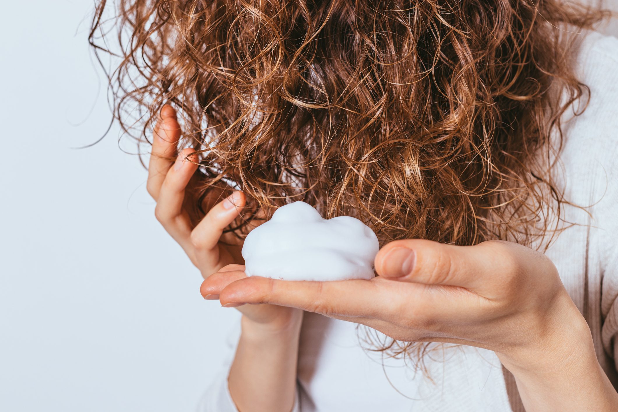 Foam rests in a woman's palm, applied to curly brown hair, against a plain white background.