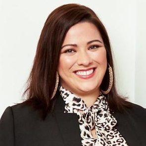 A person smiles while wearing a patterned blouse and hoop earrings, set against a plain light-colored background.