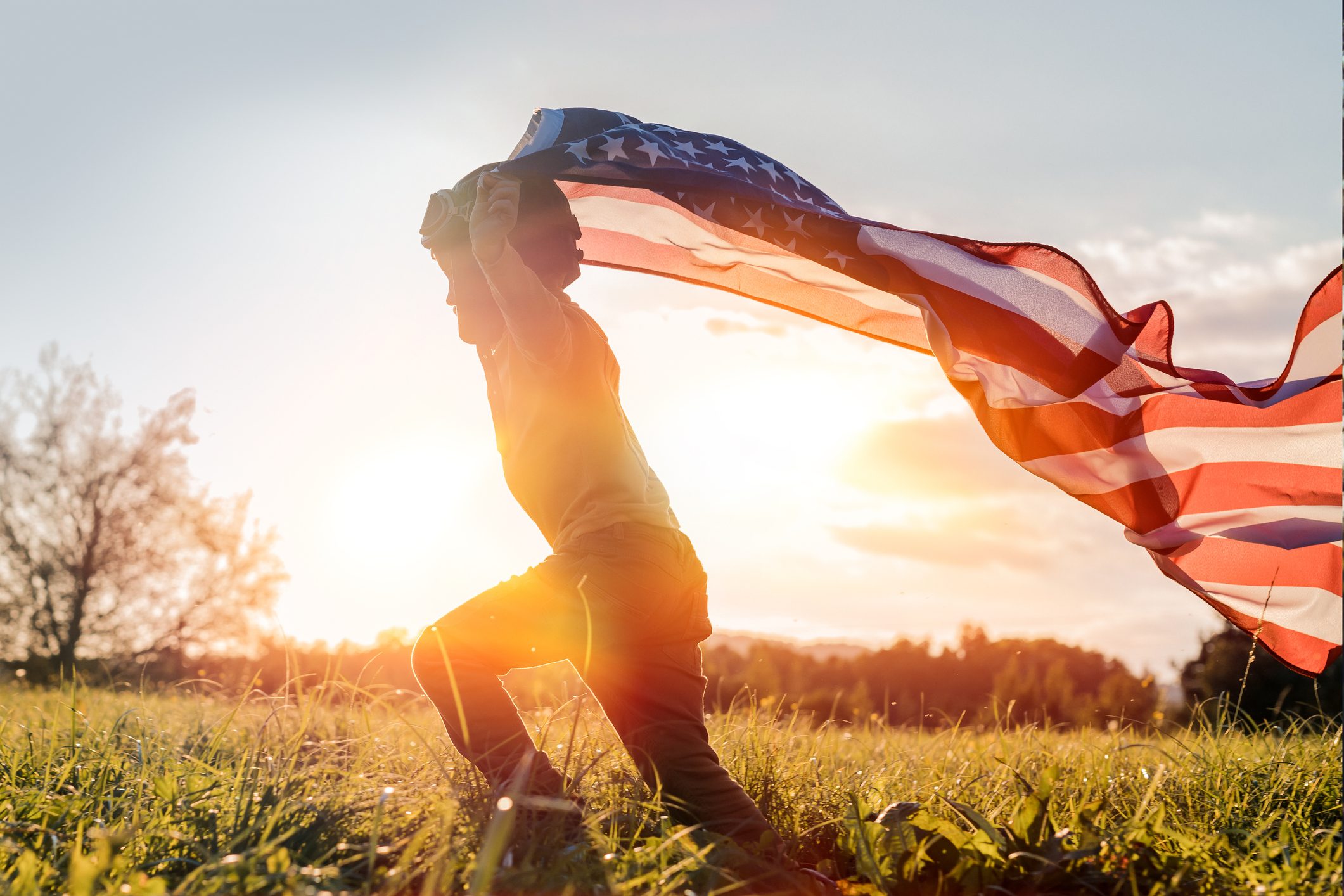 Child holds American flag, running joyfully across grassy field with the sun setting behind, creating a warm, glowing atmosphere.