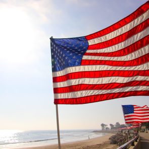 An American flag waves prominently along a sunny beach boardwalk, with more flags lining the path and palm trees in the distance.
