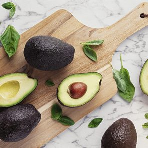 Avocados rest on a wooden cutting board surrounded by fresh basil leaves on a marble countertop.