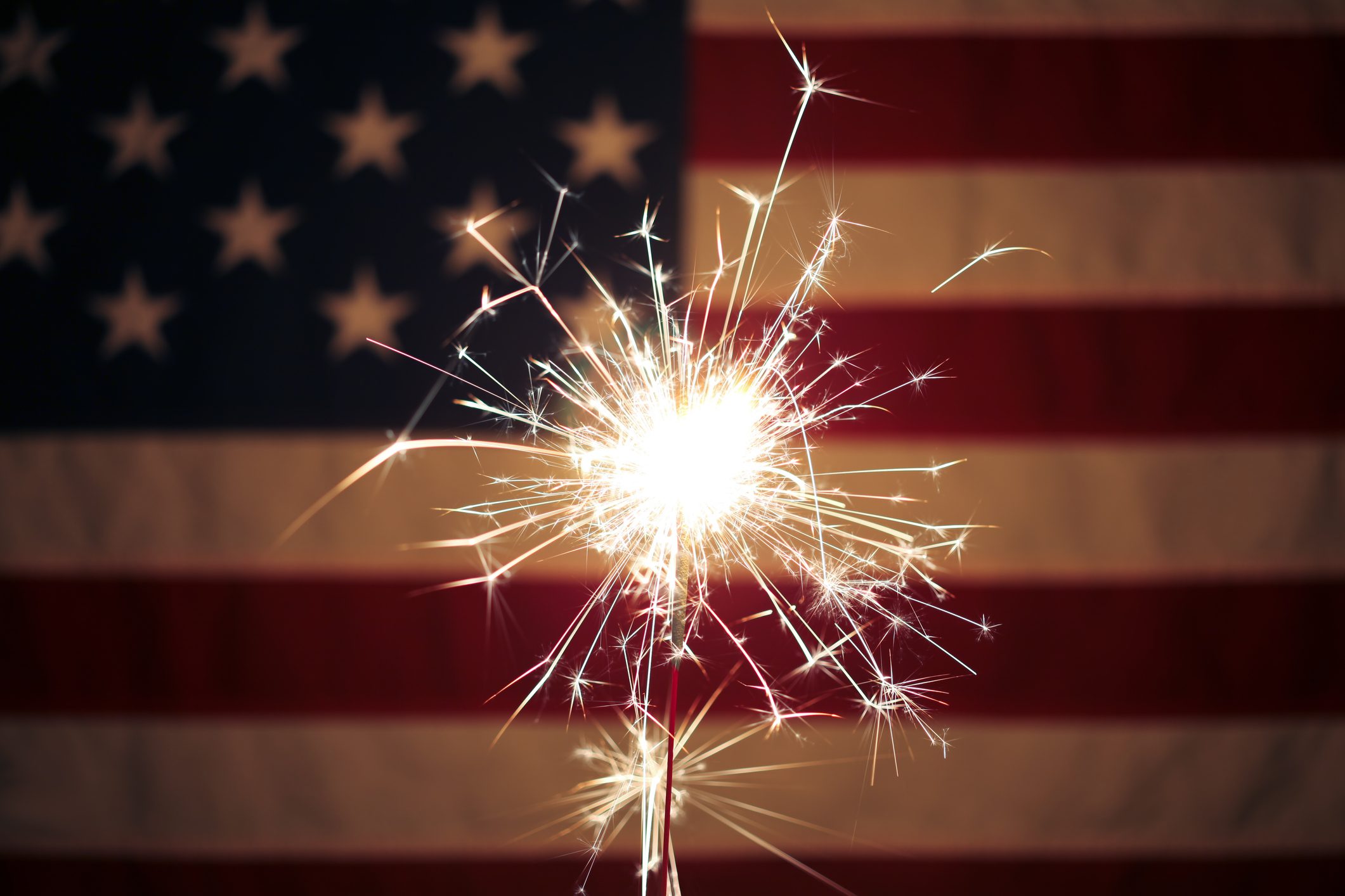 A sparkler emits bright sparks in front of an American flag backdrop, featuring visible stars and stripes.