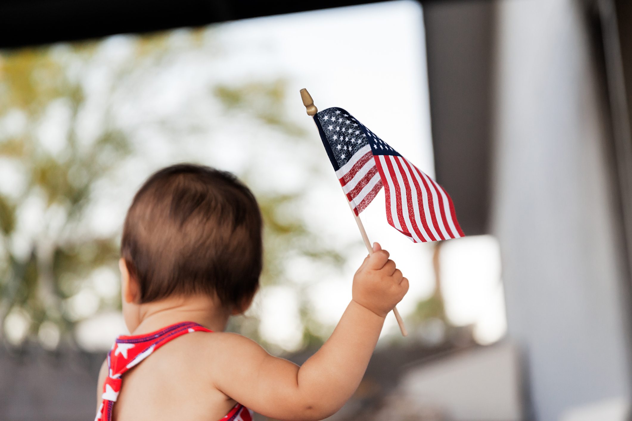 Child holding small American flag, standing outdoors with blurred trees in the background.