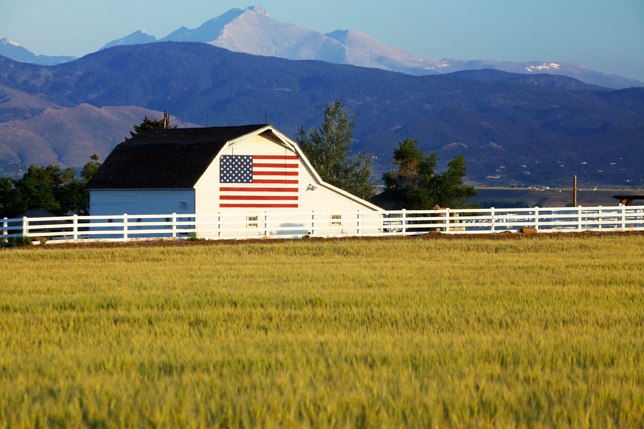 A barn displays an American flag, surrounded by a white fence and a golden wheat field, with mountains in the distant background.
