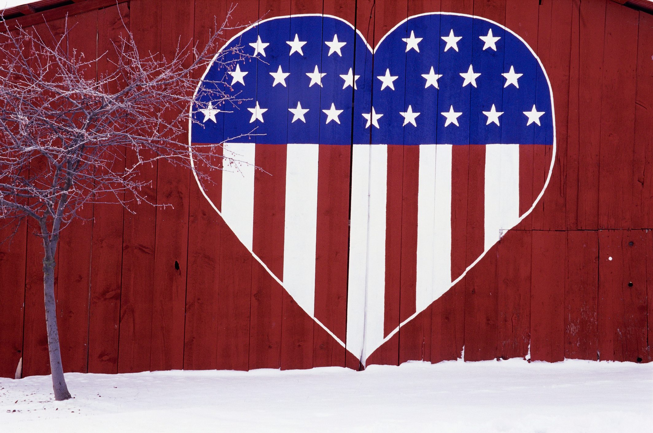 A large heart painted with the American flag design decorates a red barn wall beside a leafless tree in a snowy setting.