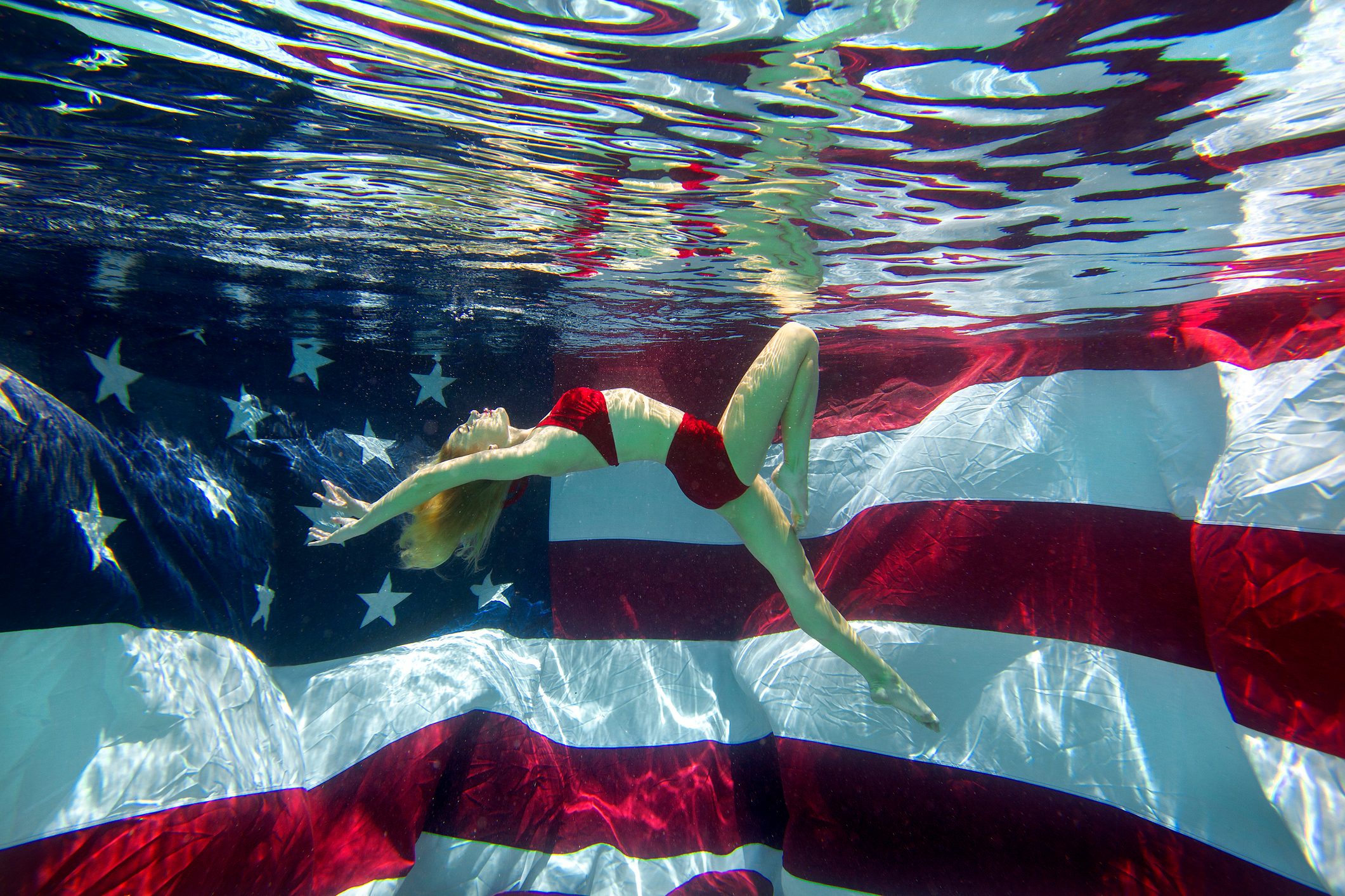 Person floating underwater, wearing a red swimsuit, against a backdrop of an American flag. The scene features rippling water effects above.