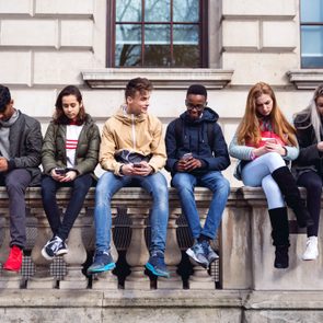 Six people sit on a stone railing, each focused on their phones, outside a building with large windows.
