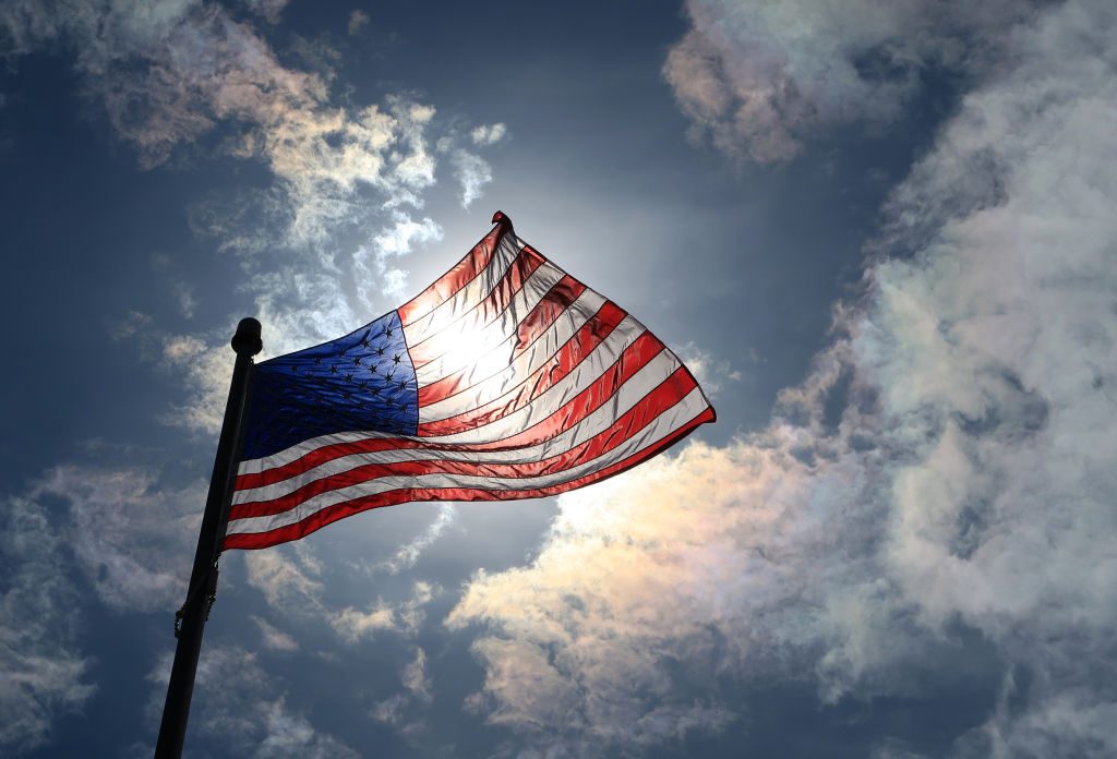 The American flag waves with sunlight shining through, surrounded by a partly cloudy sky.