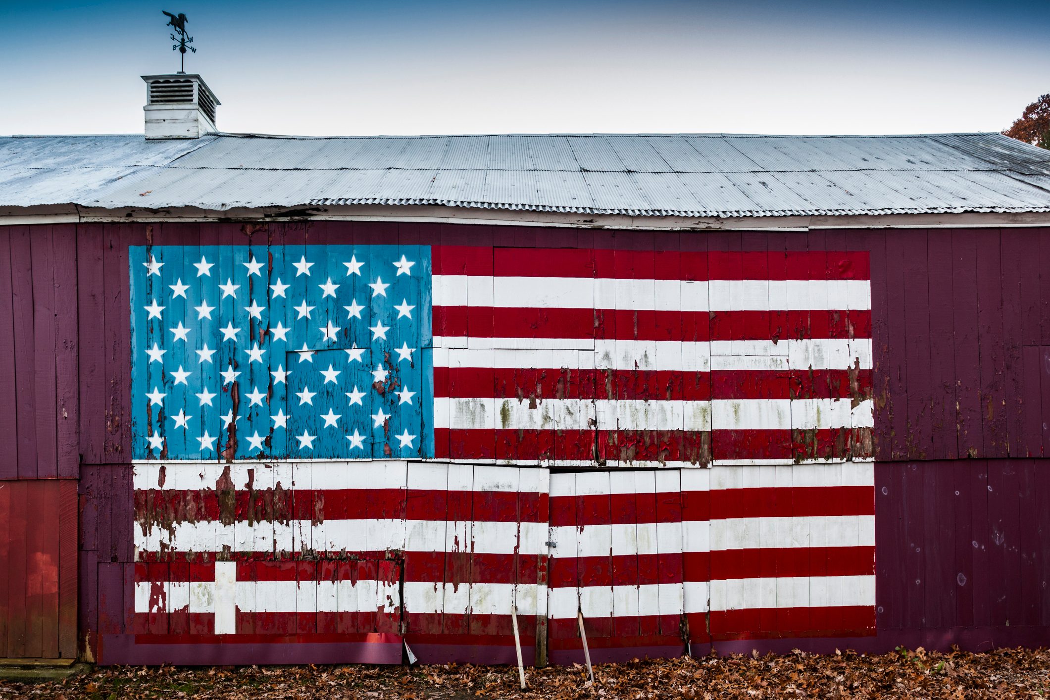 Painted American flag decorates a weathered barn wall, surrounded by fallen leaves, under a cloudy sky with a rooftop weather vane.