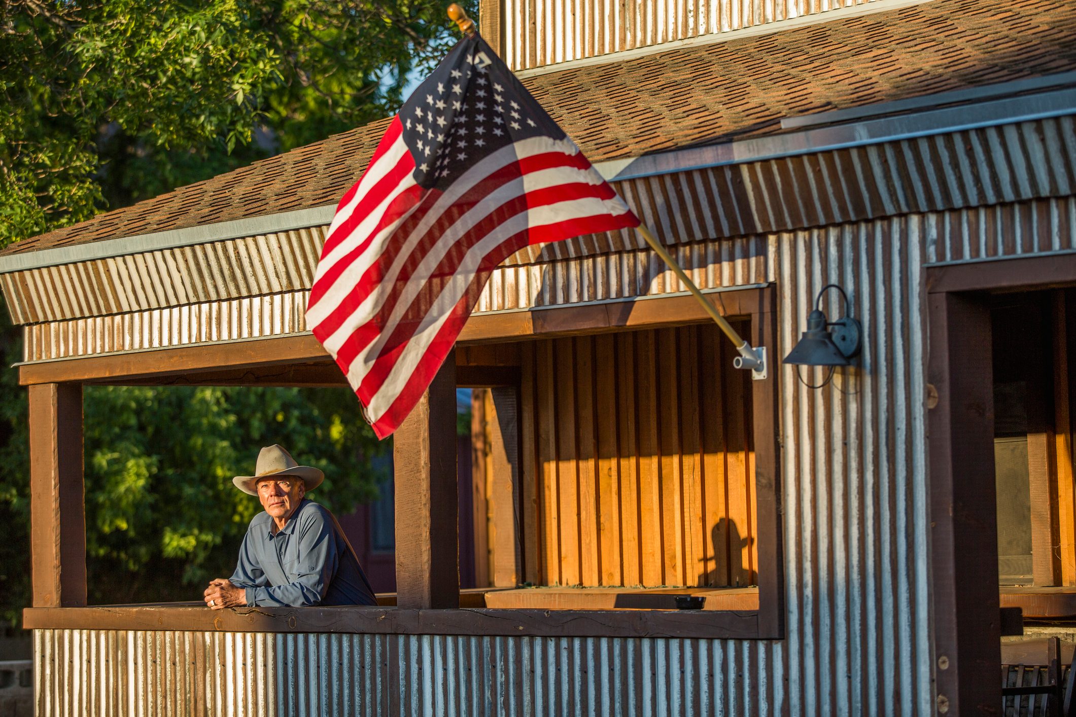 Man leans from corrugated metal porch, wearing a cowboy hat. American flag waves above him in a sunlit, rustic setting with greenery behind.