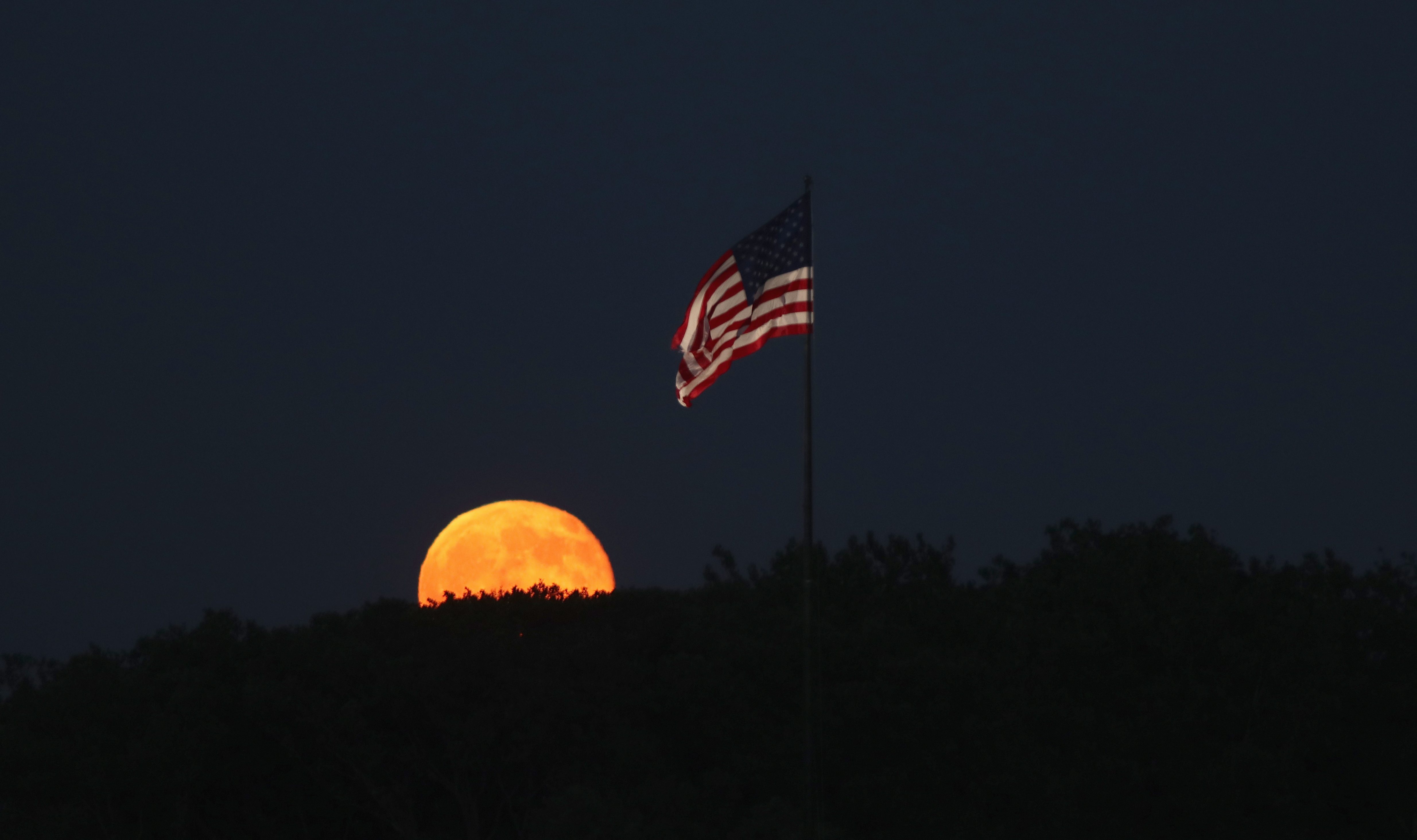 Orange moon rising over trees; American flag waves against a dark night sky.