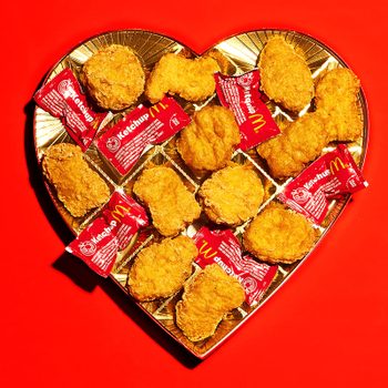 Chicken nuggets rest alongside ketchup packets on a heart-shaped golden tray, set against a vivid red background.