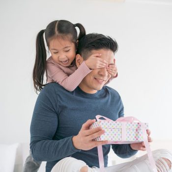Child covers seated man's eyes, both smiling brightly, in a white room. Man holds a colorful, polka-dotted gift with a pink ribbon.
