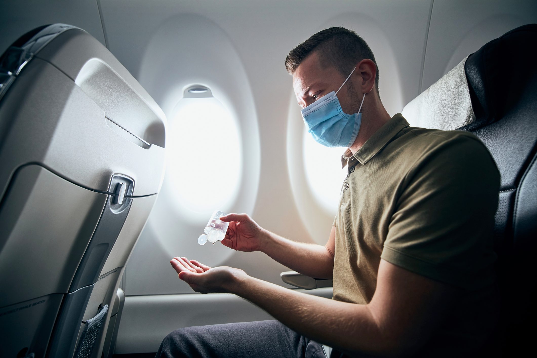 A person dispenses hand sanitizer while seated on an airplane, wearing a face mask, near a window.