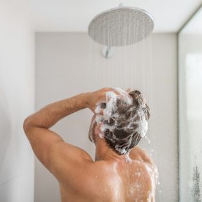 Person washing hair under shower, surrounded by foamy lather and water droplets in a tiled bathroom.