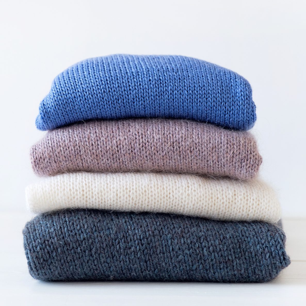 Knitted Woolen Things Of Different Colors, Stacked In A Pile, Lie On A White Wooden Table. Winter And Autumn Warm Cozy Sweaters For Charity. The Concept Of Storage, Care And Washing Of Handmade Products. Copy Of The Text Space.