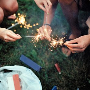 Sparklers held by hands sparkle brightly outdoors on grass, surrounded by a plastic bag and items like a lighter and boxes.