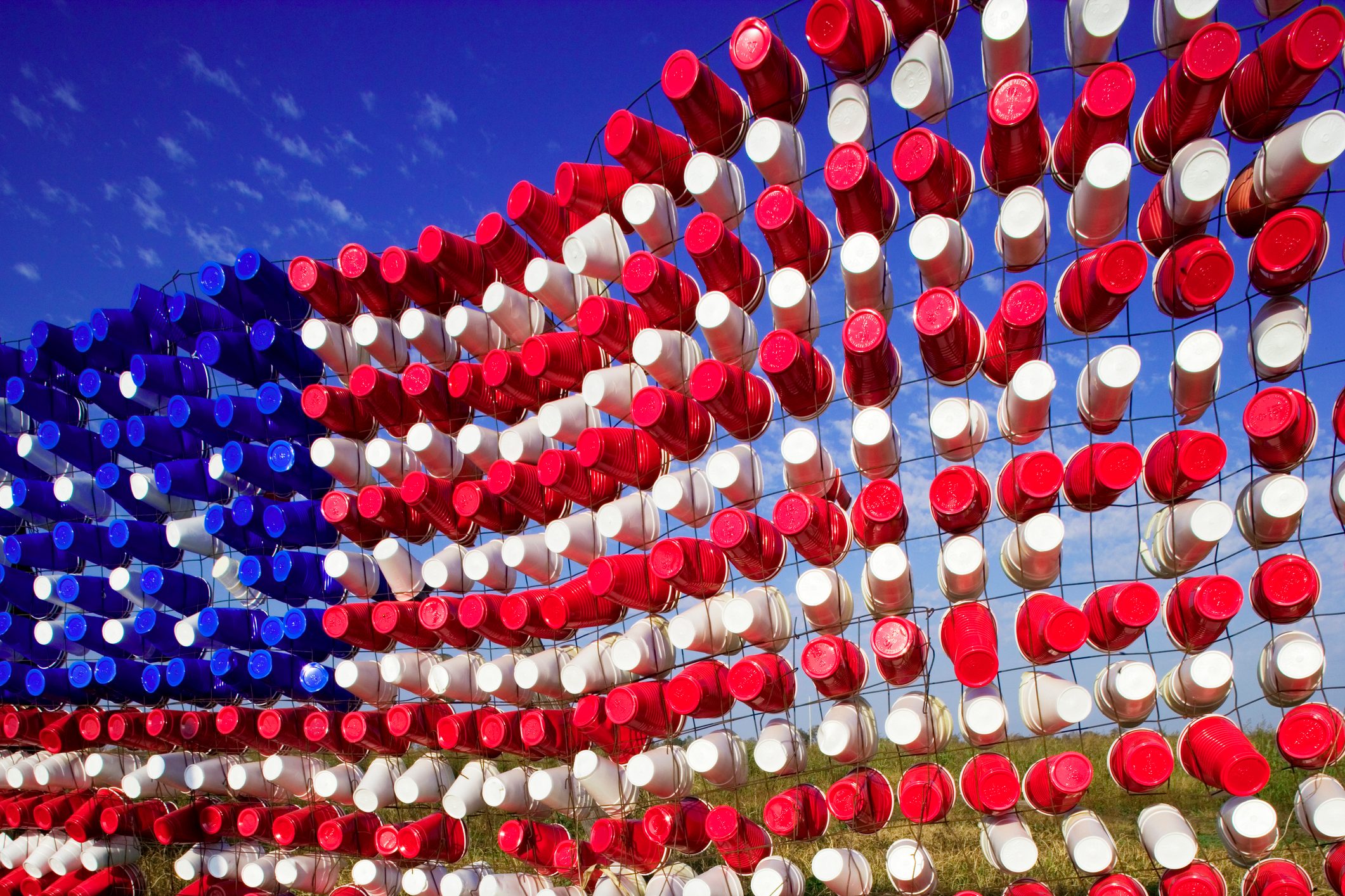 Red, white, and blue cups form a waving American flag shape against clear sky, attached to a wire grid.