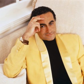 Man reclines on a sofa, wearing a yellow jacket, casually touching his forehead. The setting is a bright, comfortable living room.