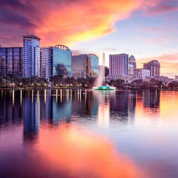 Fountain sprays water, reflected in a tranquil lake, surrounded by city skyscrapers under a dramatic orange and pink sunset sky.