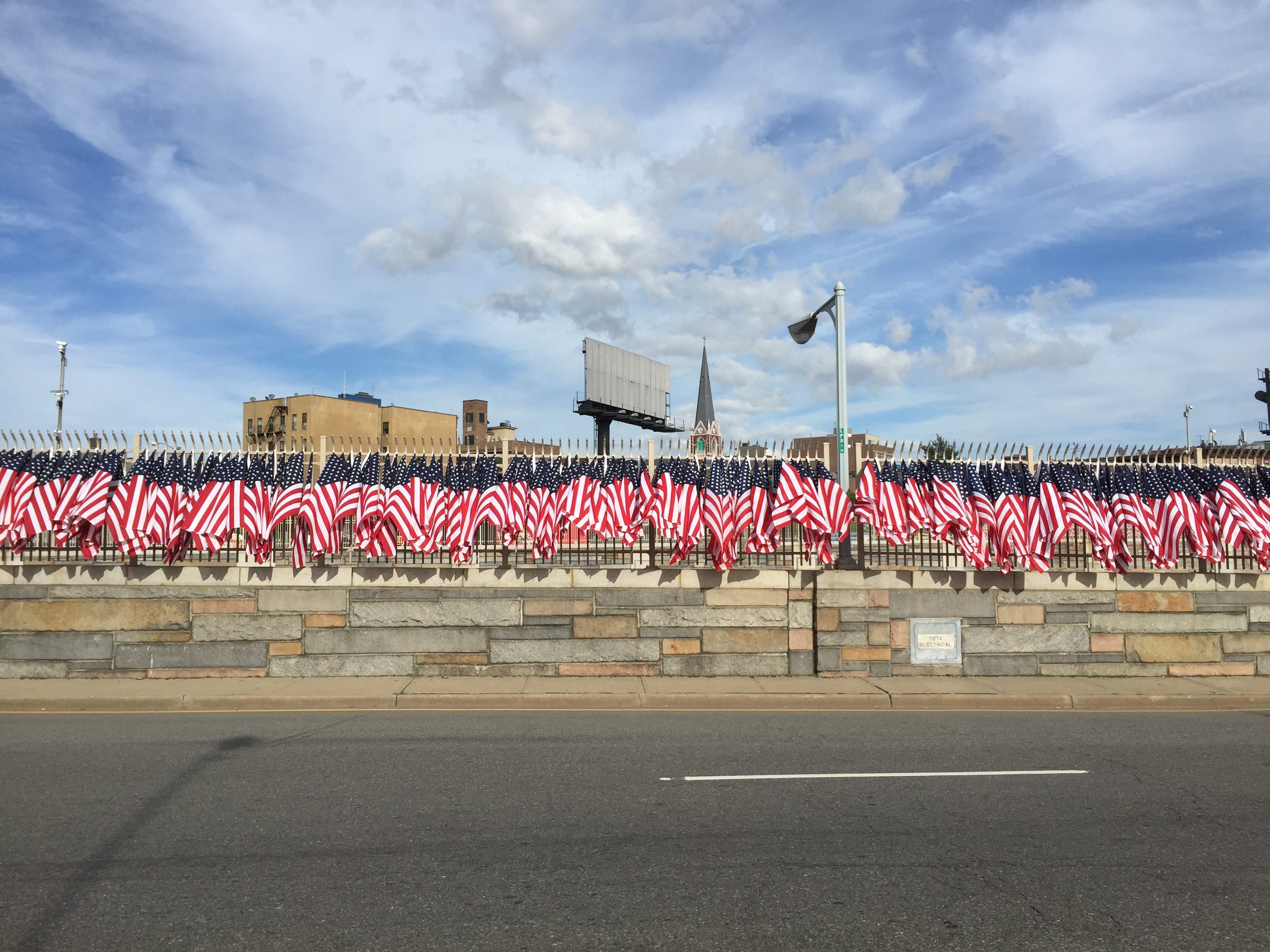 American flags wave on a long fence bordering a stone wall by a road, under a partly cloudy blue sky in an urban area.