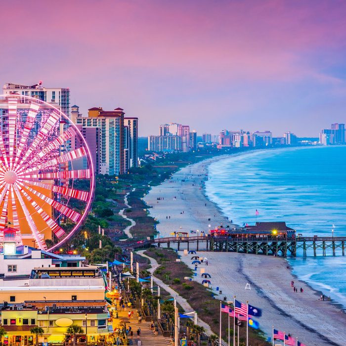 Ferris wheel illuminated, spins near beachside boardwalk, surrounded by high-rise buildings at sunset with ocean waves gently washing ashore.