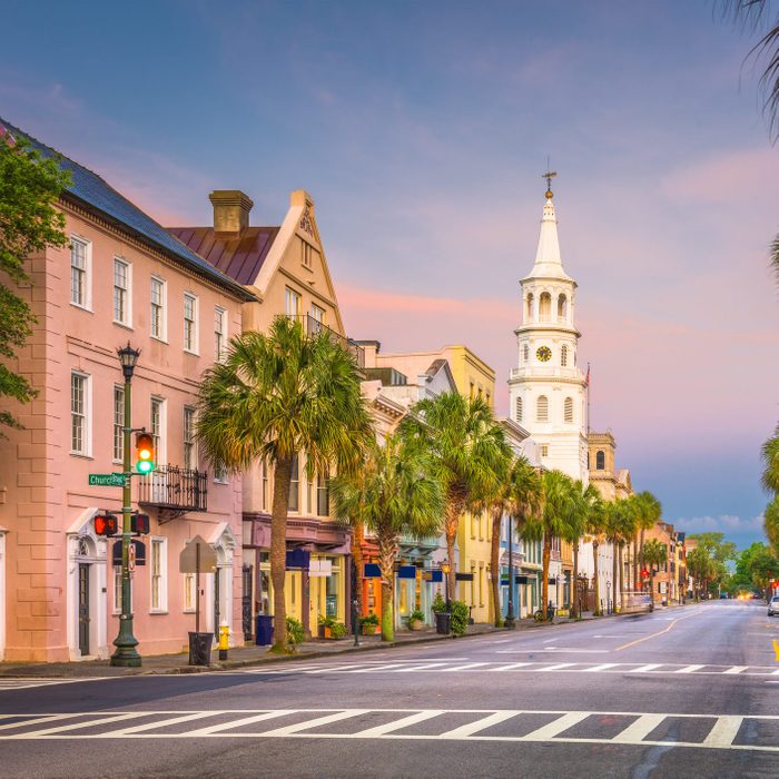 Street lined with colorful historic buildings and palm trees; a white church steeple rises in the distance. Traffic signal glows green, sign reads 