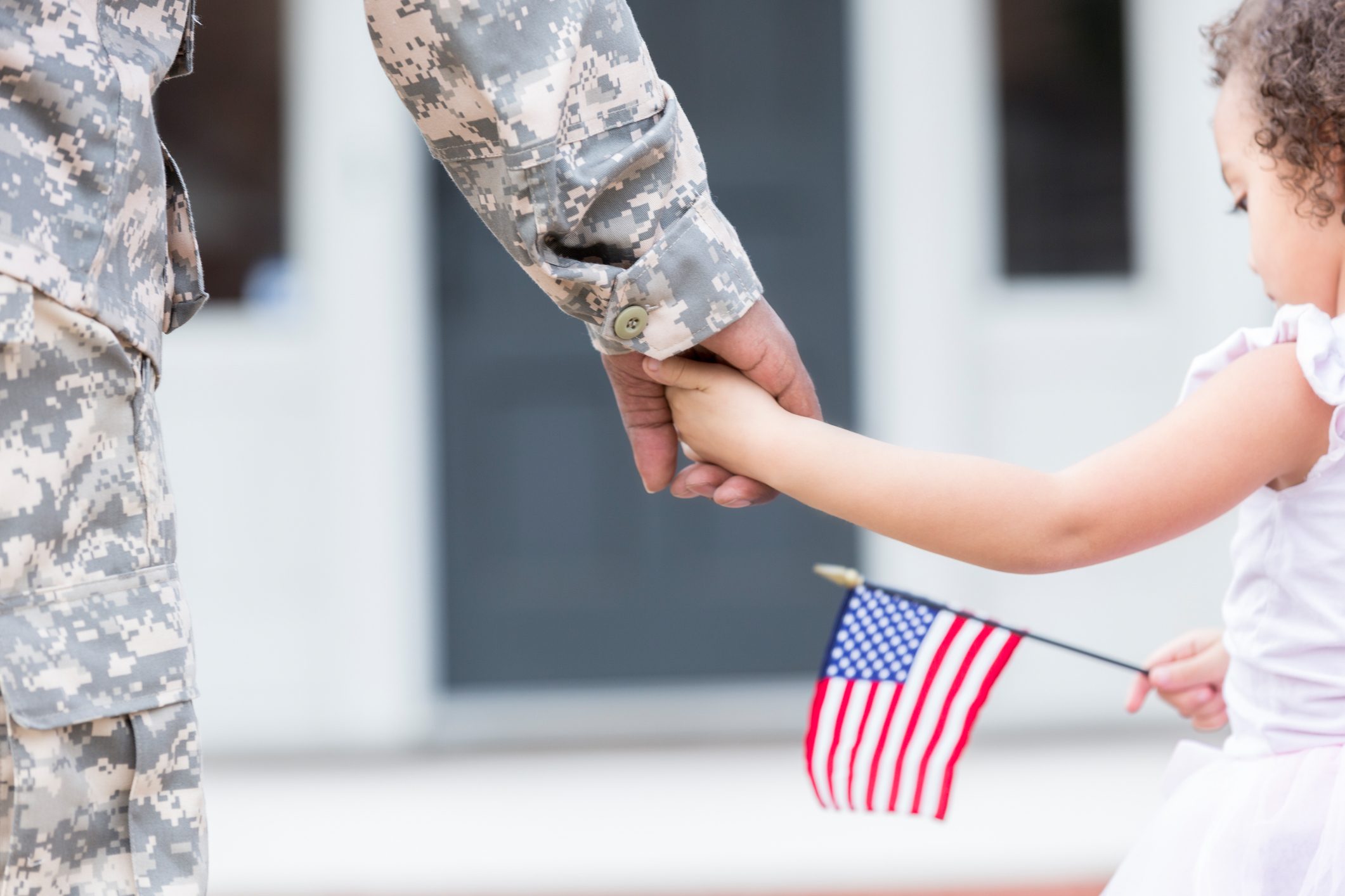 A soldier in camouflage holds a child's hand. The child is holding a small American flag, outdoors near a building.