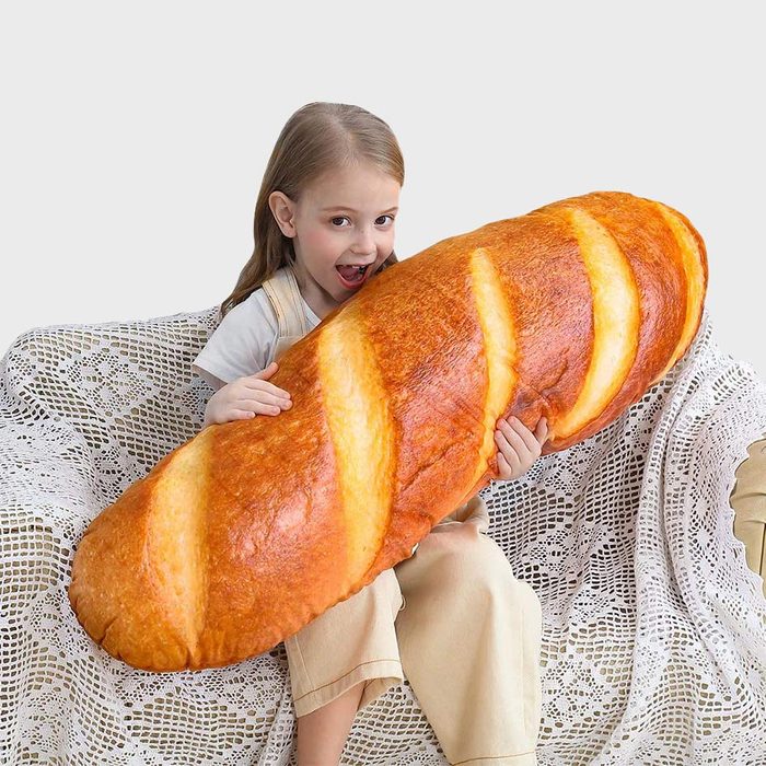 A child cuddles a giant bread-shaped pillow, sitting on a lacy, white-covered sofa against a plain background.