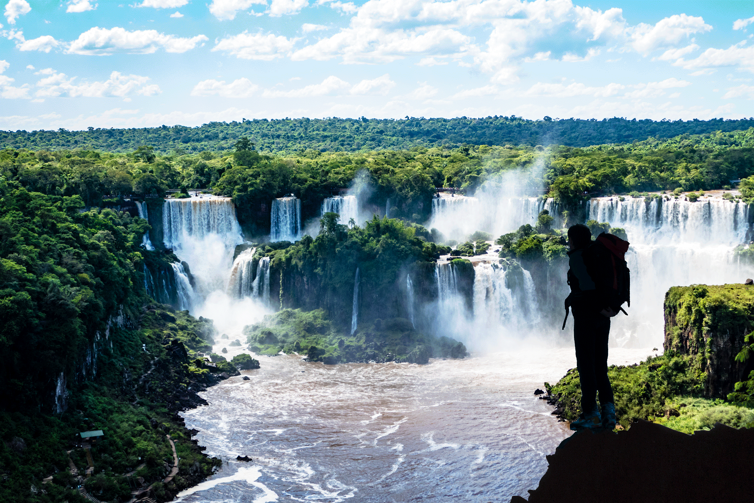 A person stands overlooking waterfalls, streaming amidst lush green forest under a partly cloudy sky.