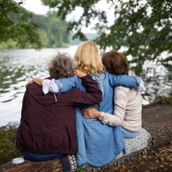 Three people sit on a log, embracing each other, by a serene lake surrounded by greenery.