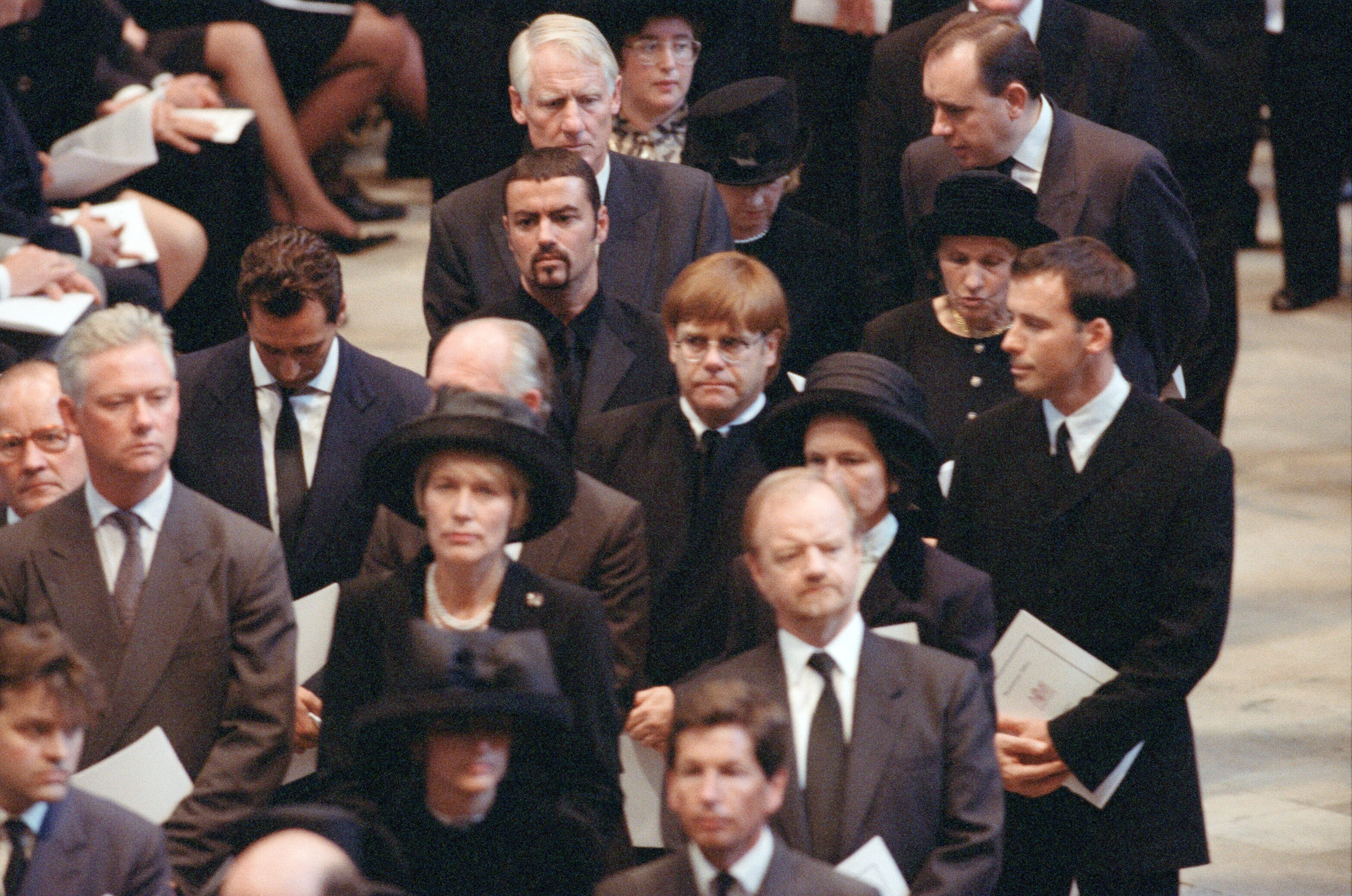 People standing solemnly, holding programs, in a formal indoor setting, suggestive of a ceremony or memorial event.