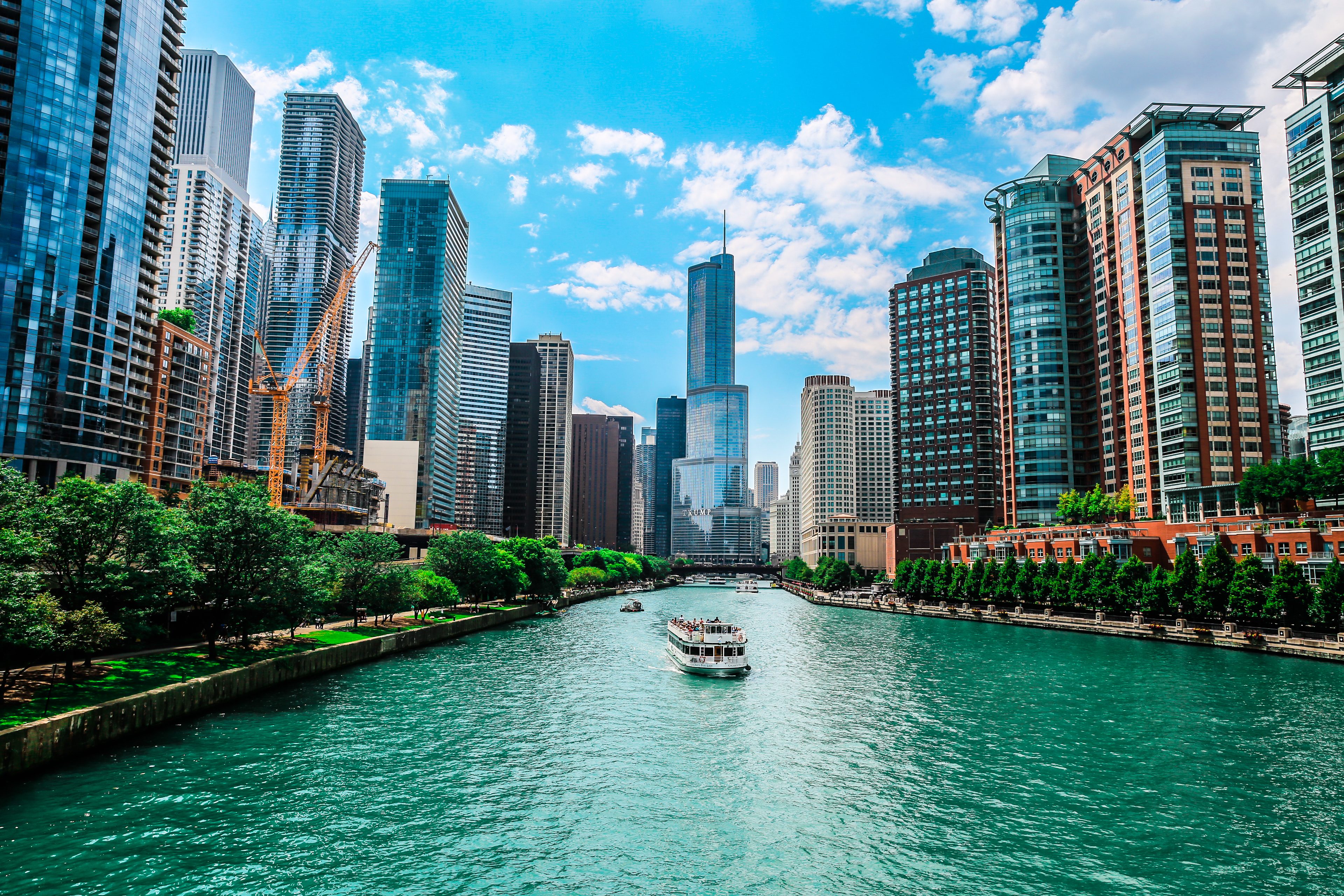 Boat sails through river, surrounded by skyscrapers and lush greenery under a bright blue sky.