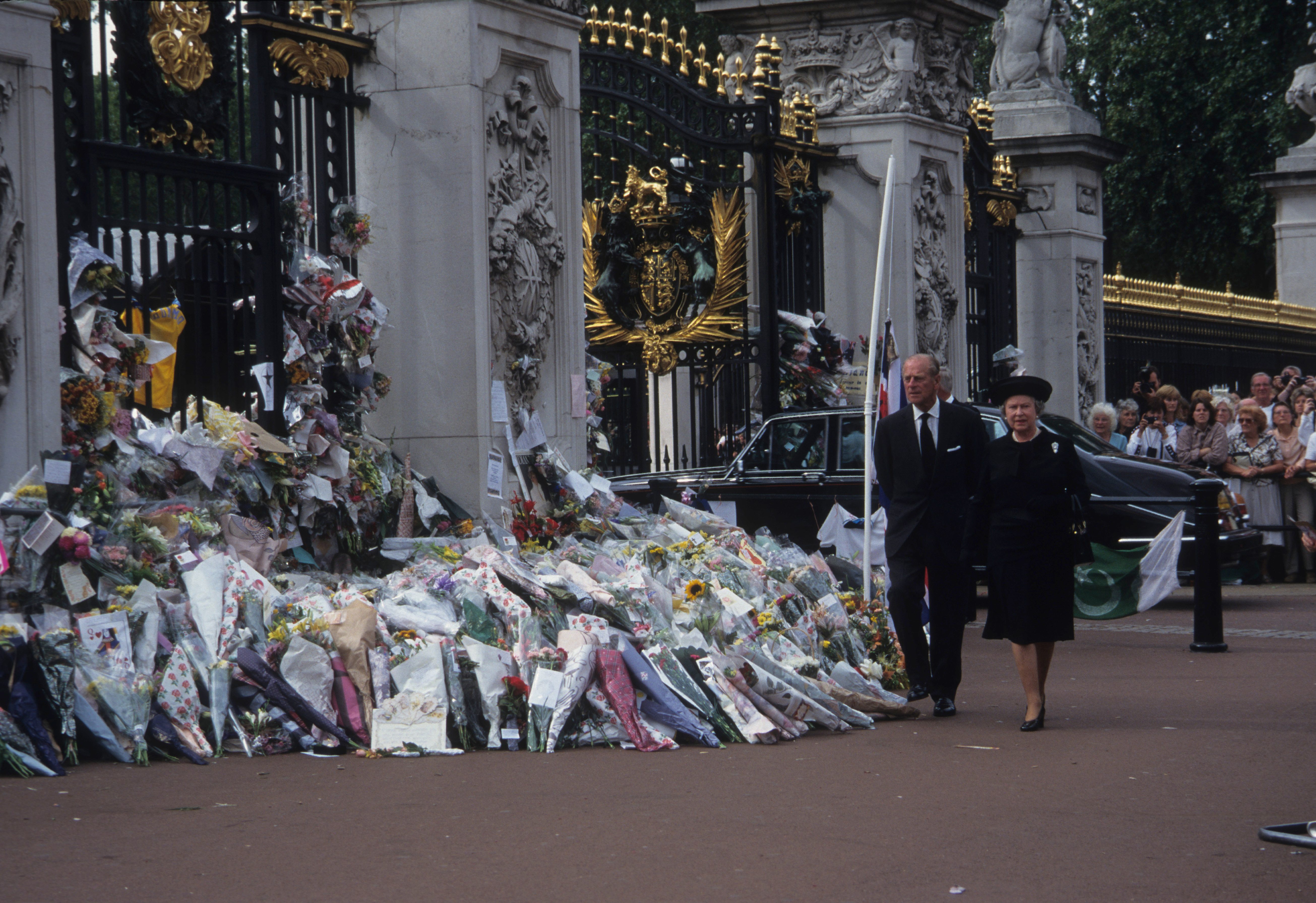 Flowers lay in tribute outside ornate gates; two individuals in formal attire walk past, with a crowd and a parked car visible nearby.
