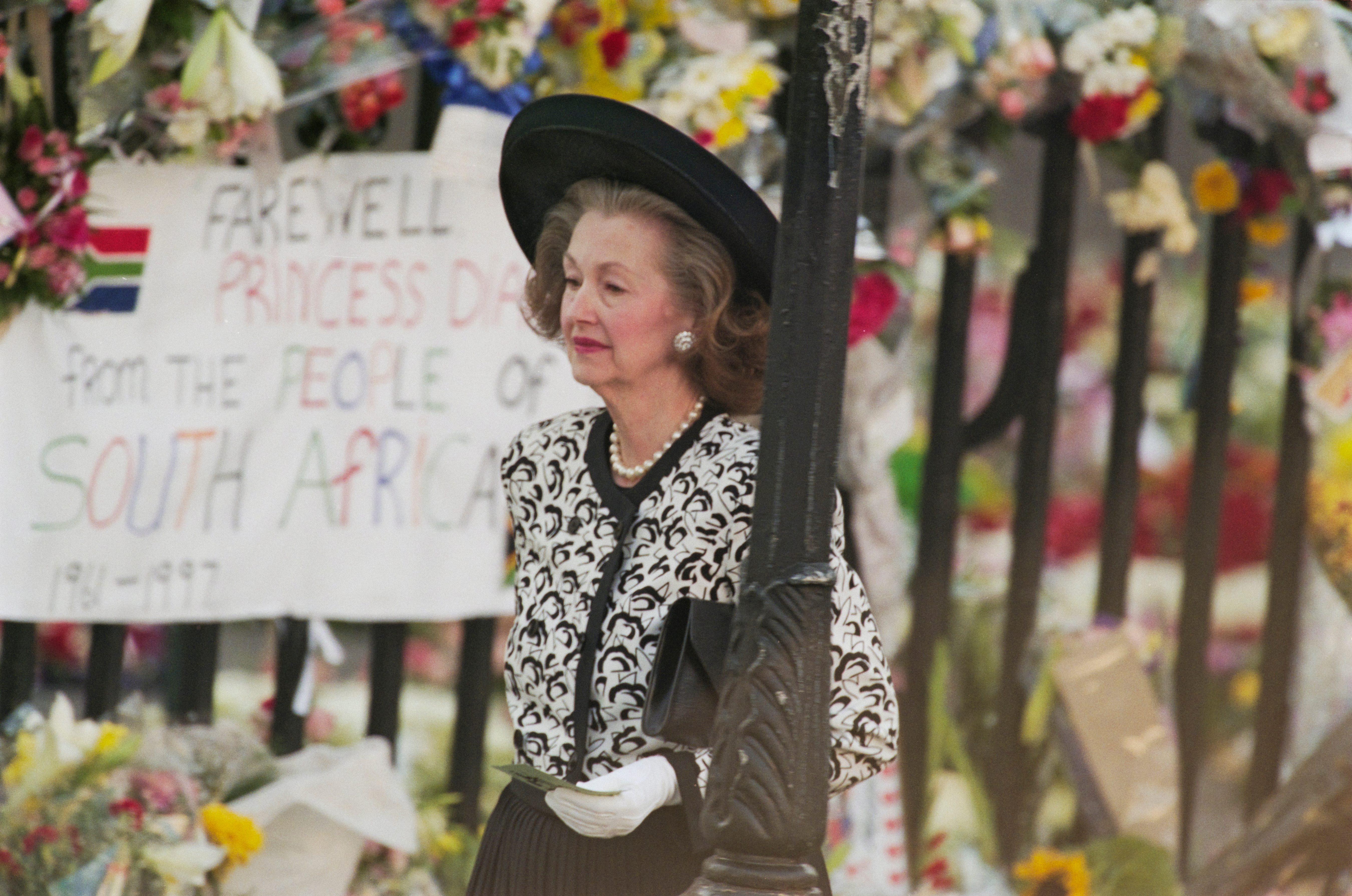 A woman stands looking contemplative, dressed in formal attire, holding a card, in front of a floral memorial with a tribute to Princess Diana.