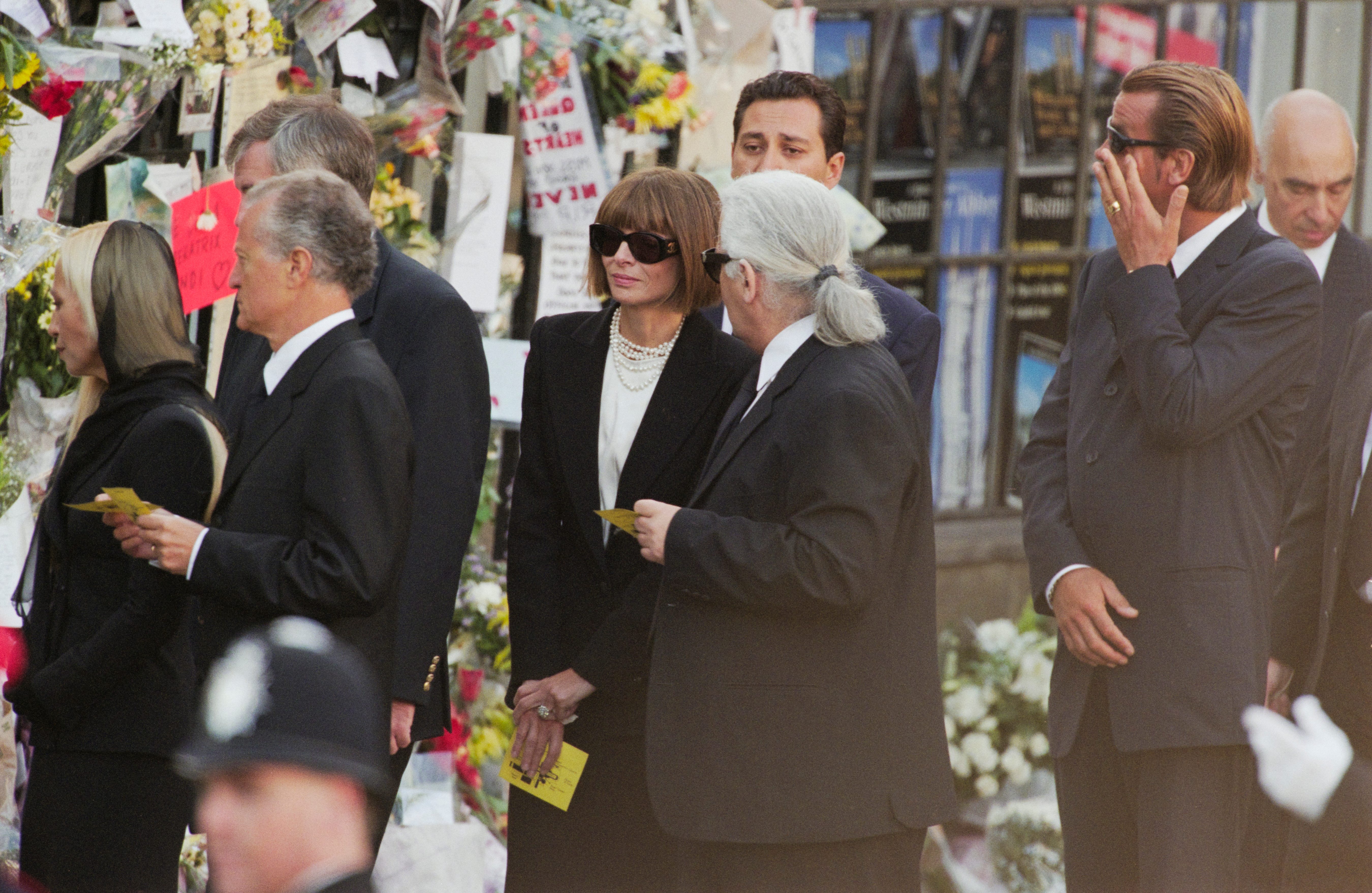 People in formal attire gather, holding papers, near a wall covered with flowers and notes. A police officer's helmet is visible in the foreground.