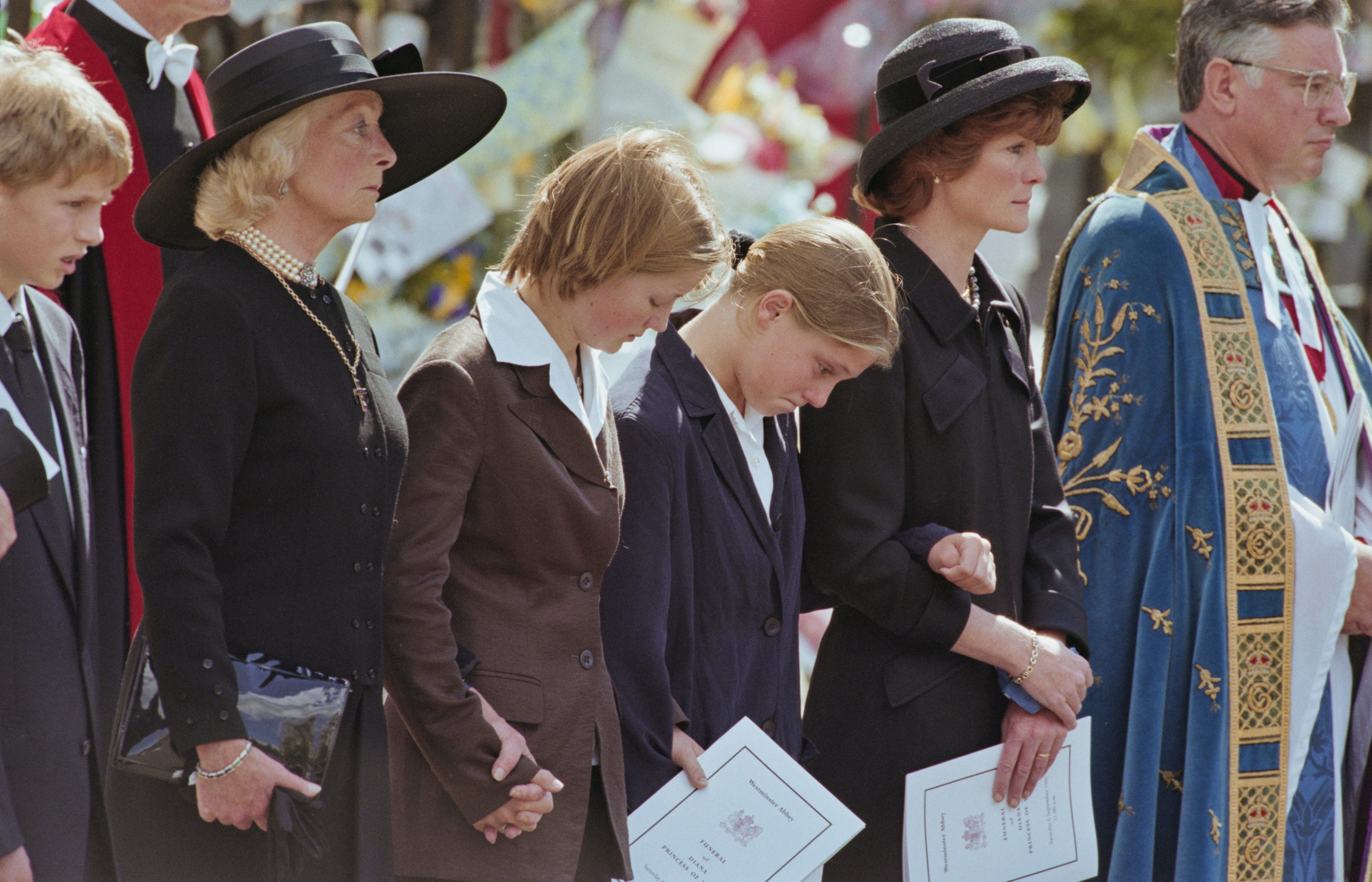 Mourners stand solemnly holding programs, dressed in dark formal attire, during an outdoor memorial service surrounded by flowers. Programs read: "Funeral Service of Diana, Princess of Wales."