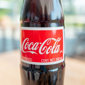 A Coca-Cola bottle stands upright on a blurred, outdoor table. Text: 