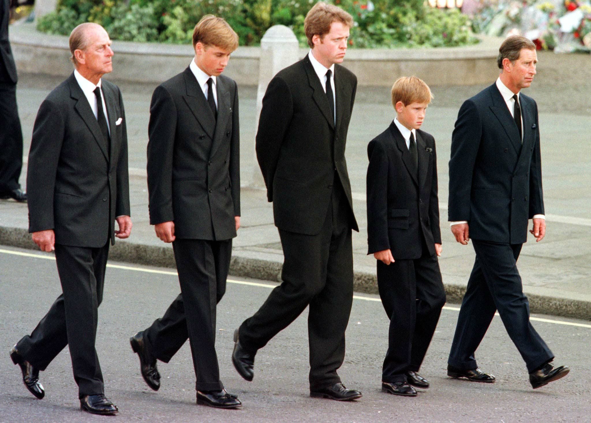 Five men in black suits walk somberly on a city street, surrounded by greenery and flowers in the background.