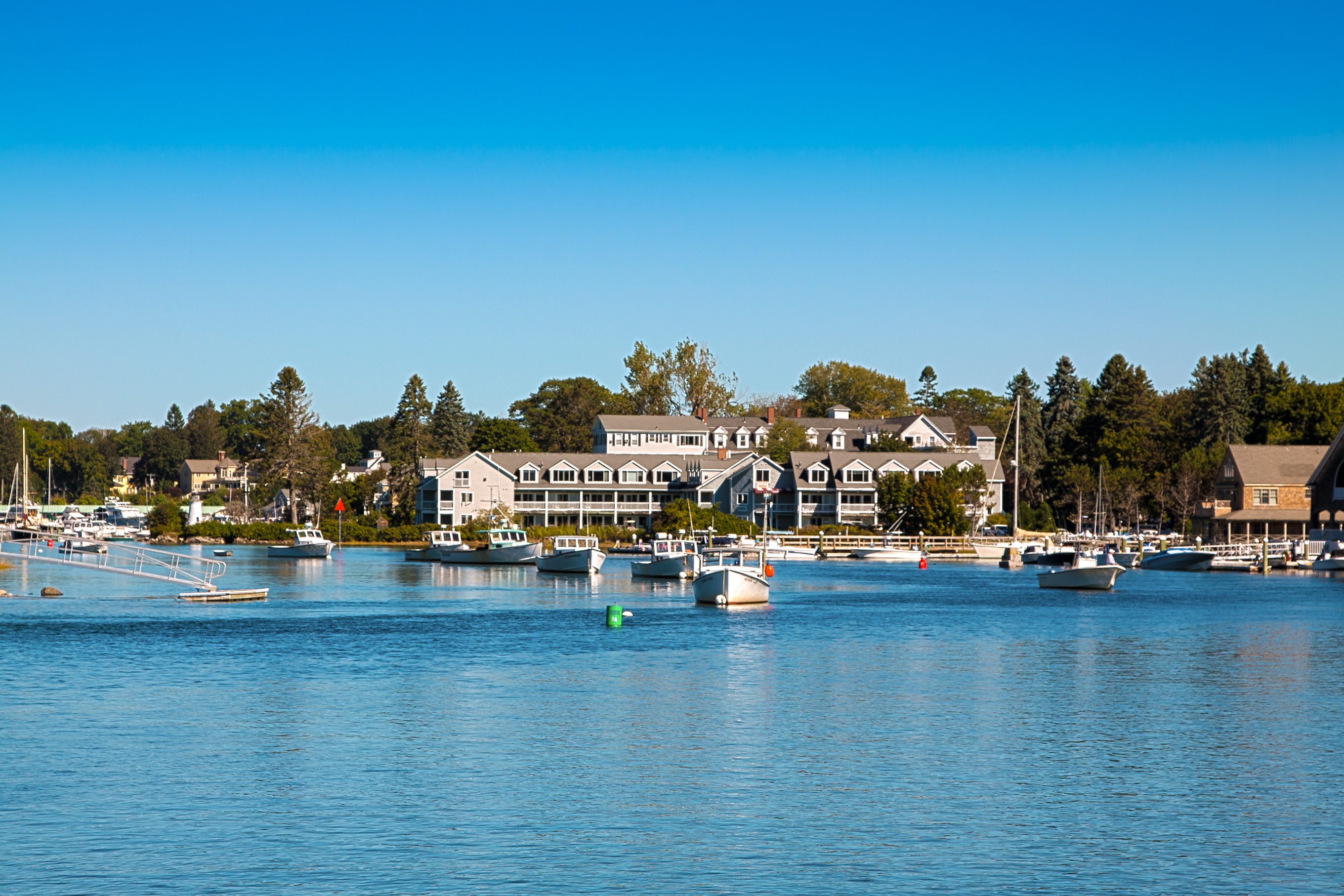 Boats float in a calm harbor, surrounded by quaint buildings and green trees in clear weather.
