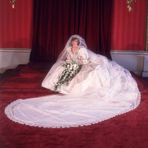 A bride sits gracefully with a long white gown and floral bouquet, surrounded by elegant red drapery and plush carpet.