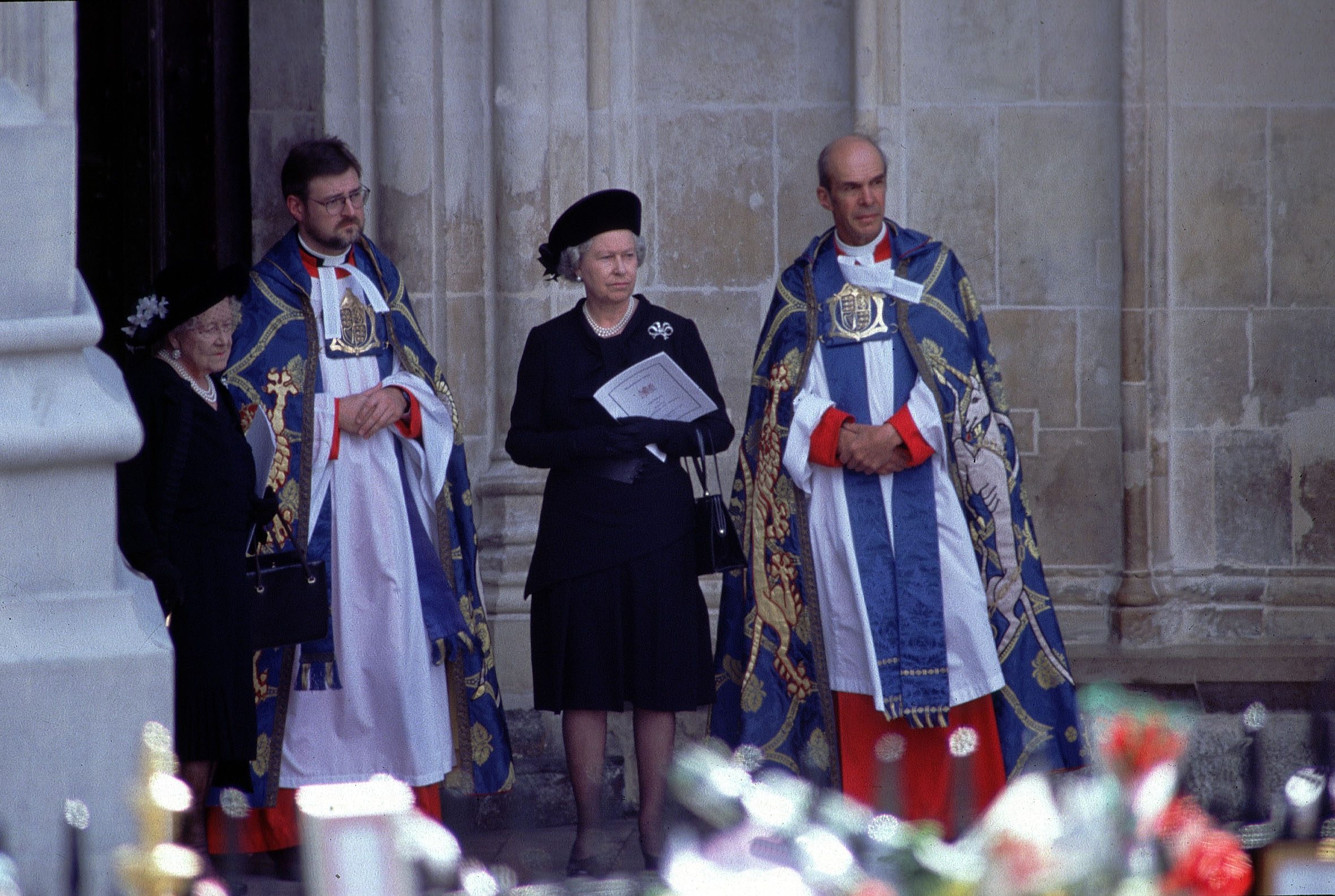 Clergy in ornate robes stand solemnly with two women in black dresses and hats, holding papers, outside a stone building, with flowers in the foreground.