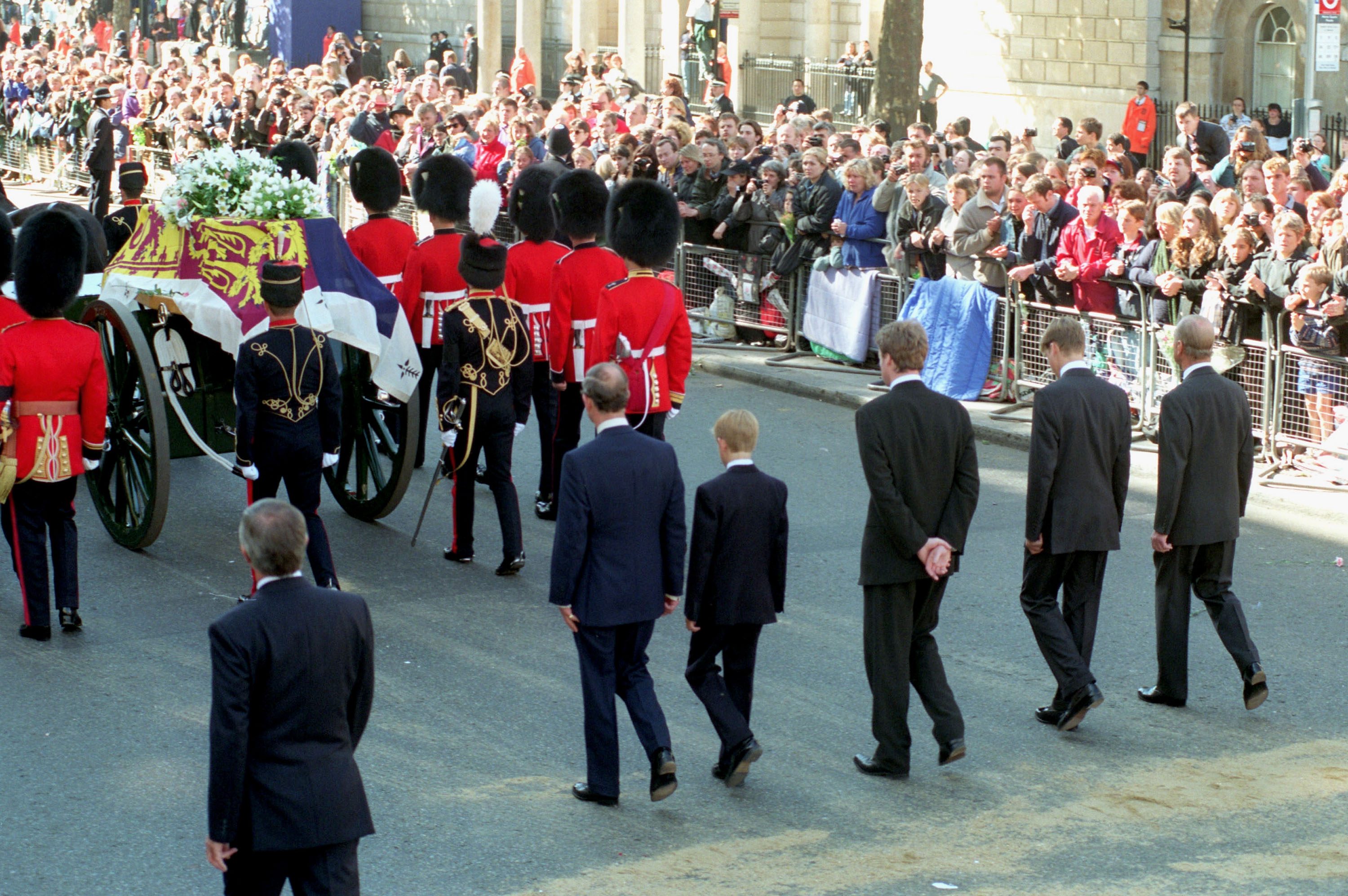 Casket draped with a flag, adorned with flowers, is escorted by guards and men in suits, with a crowd observing from behind barricades.