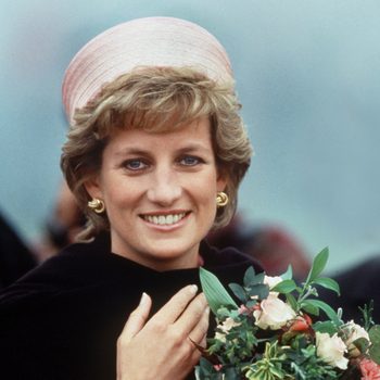 A person smiles while holding a bouquet of flowers, wearing a pink hat and earrings, set against a blurred outdoor background.